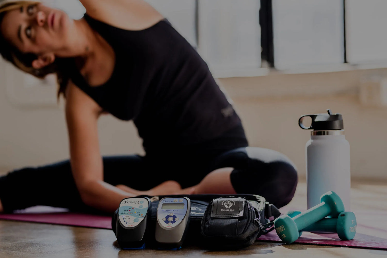 woman doing a yoga position on a workout mat with biomodulator pro and plus, small and large carrying case, hand weights and water bottle